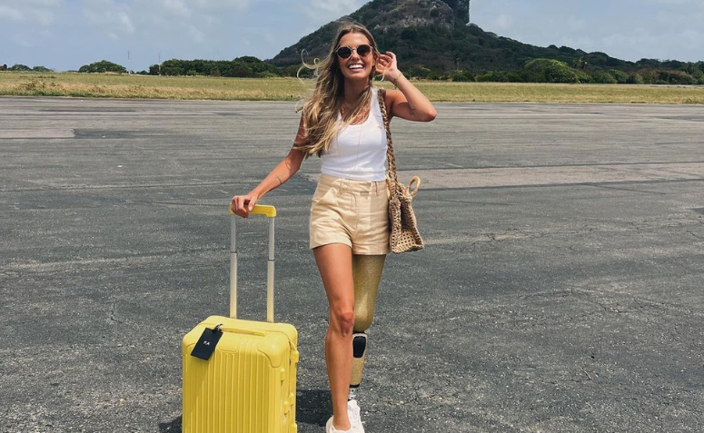 Mulher com cabelos longos e loiros, vestindo blusinha branca e short amarelo, está com uma mala também amarela